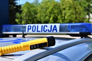 Dach policyjnego radiowozu oznakowanego. Na środku dachu umieszczona belka z napisem POLICJA, przed nia położone urządzenie do badania stanu trzeźwości. W tle drzewa.