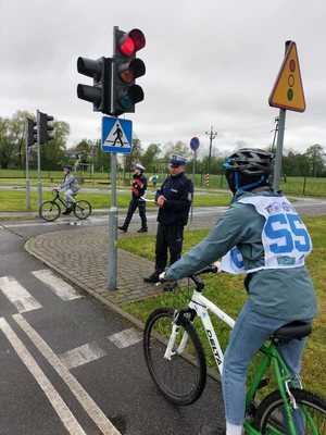 Dziecko na rowerze stojące przed sygnalizatorem wysyłającym sygnał czerwony. Obok umundurowany policjant.