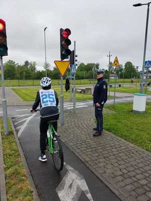 Dziecko na rowerze stojące przed sygnalizatorem wysyłającym sygnał czerwony. Obok umundurowany policjant.