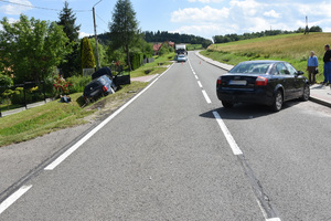 Na zdjęciu droga asfaltowa. Po lewej stronie, w rowie leżący samochód marki audi. Na prawym pasie pojazd marki audi stojący w poprzek jezdni. Obok tego pojazdu dwaj stojący mężczyźni, rozmawiają ze sobą. Jeden z nich ma zabandażowaną rękę. W tle inne pojazdy oraz zabudowania.