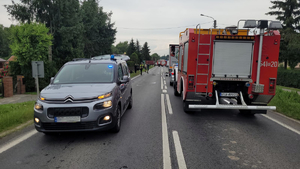 Droga asfaltowa. Na prawym pasie stojący samochód straży pożarnej. Widać auto od tyłu. Na lewym pasie nieoznakowany radiowóz z włączonym sygnałem świetlnym barwy niebieskiej. W tle zabudowania oraz drzewa.