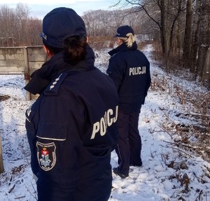 Dwie umundurowane policjantki kontrolujące pustostany pod kątem ujawniania osób bezdomnych. W tle drzewa i pozostałości murów. Na trawie leży śnieg.