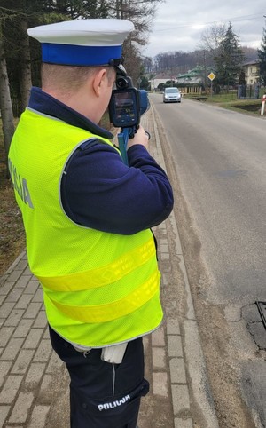 Policjant Ruchu Drogowego prowadzący działania Prędkość- Trzyma w ręce urządzenie do pomiaru prędkości i tarczę służącą do zatrzymywania pojazdów. Zatrzymuje do kontroli żółty samochód osobowy.