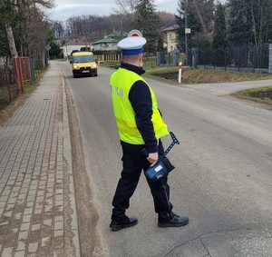 Policjant Ruchu Drogowego prowadzący działania Prędkość- Trzyma w ręce urządzenie do pomiaru prędkości i tarczę służącą do zatrzymywania pojazdów. Zatrzymuje do kontroli żółty samochód osobowy.