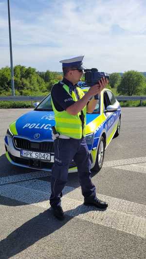 Umundurowany policjant trzyma w ręce miernik prędkości.  Tuz za nim widać oznakowany radiowóz policyjny.