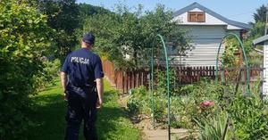 Umundurowany policjant kontrolujący bezpieczeństwo w Rodzinnych Ogródkach Działkowych. W tle domek letniskowy.