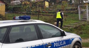 Umundurowany policjant kontrolujący tereny Rodzinnych Ogródków Działkowych. Na zdjęciu widoczny jest również oznakowany radiowóz.