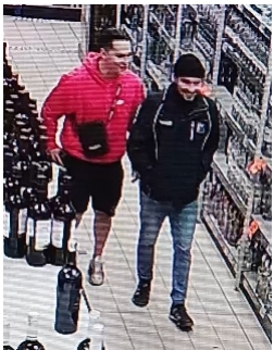 Na zdjęciu dwaj mężczyźni. Jeden ubrany był w czerwoną bluzę z kapturem, czarne krótkie spodenki, sportowe buty. Miał również przy sobie męską czarną torebkę, saszetkę. Drugi z mężczyzn na głowie miał czarną czapkę, ubrany był w czarną kurtkę z białymi elementami, niebieskie jeansowe spodnie oraz czarne sportowe buty. W tle sklepowe regały.