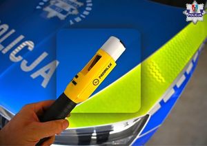 Urządzenie do badania stanu trzeźwości trzymane w ręce przez umundurowanego policjanta. W tle radiowóz oznakowany.
