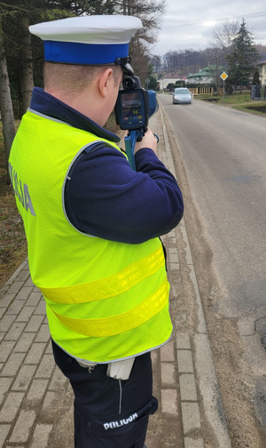 Umundurowany policjant stojący na poboczu drogi, używający urządzenia do mierzenia prędkości pojazdów poruszających się po drodze.
