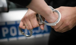 Na zdjęciu widać dłonie ludzkie. Na jedną są zakładane kajdanki. w tle napis policja  na drzwiach radiowozu,