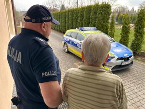 Umundurowany policjant rozmawiający ze starszą kobietą. w tle oznakowany radiowóz.