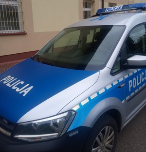 Radiowóz oznakowany w deszczu. Samochód jest zaparkowany przy budynku o żółtych ścianach.