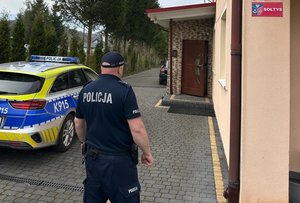 Umundurowany policjant idący w stronę drzwi do domu. Na ścianie budynku czerwona, prostokątna tabliczka z białym napisem SOŁTYS. Po lewej stronie zaparkowany oznakowany radiowóz. W tle drzewa iglaste.