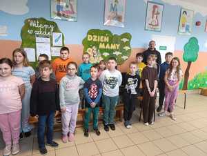 Na zdjęciu grupa dzieci w wieku szkolnym, w tle umundurowany policjant. Zdjęcie wykonane w budynku szkoły.