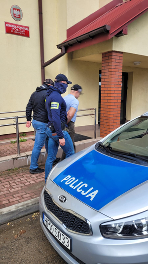 Policjanci prowadzą mężczyznę, który został zatrzymany. Ma on założone kajdanki i wchodzi razem z policjantami do żółtego budynku. Na zdjęciu jest widoczny oznakowany radiowóz.