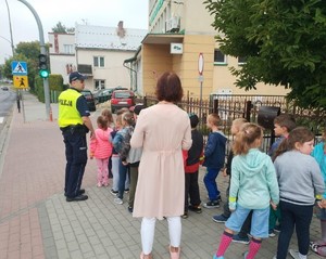 Policjant uczący grupę dzieci jak prawidłowo zachować się  przy przechodzeniu przez jezdnię.