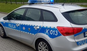 Na zdjęciu oznakowany radiowóz policyjny. W tle fragment betonowego ogrodzenia i trawnik.