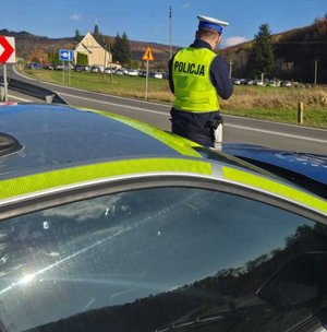 Policjant pracujący przy drodze. W tle widoczne zabudowania i fragment drogi.