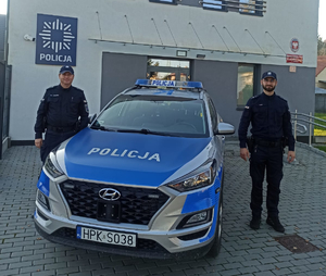 Dwaj umundurowani policjanci stojący przy oznakowanym radiowozie. W tle budynek w kolorze szarym.