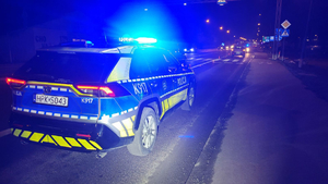 Zdjęcie z miejsca zdarzenia. Na fotografii radiowóz oznakowany z włączonymi światłami błyskowymi w kolorze niebieskim. W tle droga oraz pojazdy. Widoczny uszkodzony pojazd. Zdjęcie wykonane nocą, jest ciemno.