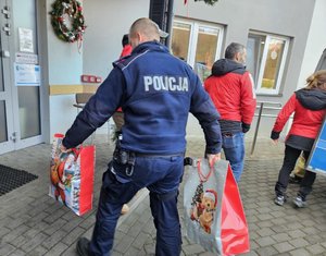Umundurowani policjanci noszący paczki mikołajowe dla podopiecznych DPSu.  Policjanci biorą paczki z samochodu i wnoszą do budynku, w którym odbędzie się wręczanie prezentów autorom listów.