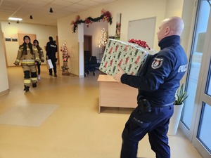 Umundurowani policjanci noszący paczki mikołajowe dla podopiecznych DPSu.  Policjanci biorą paczki z samochodu i wnoszą do budynku, w którym odbędzie się wręczanie prezentów autorom listów.