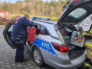 Umundurowani policjanci noszący paczki mikołajowe dla podopiecznych DPSu.  Policjanci biorą paczki z samochodu i wnoszą do budynku, w którym odbędzie się wręczanie prezentów autorom listów.
