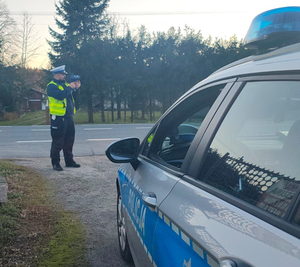 Na zdjęciu, na pierwszym planie widać fragment radiowozu oznakowanego. Na drugim stojącego umundurowanego policjanta, który dba o bezpieczeństwo i porządek na drodze.