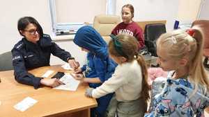 Dzieci stojące przy biurku. Policjantka przy użyciu tuszu wykonuje na kartce odbicia odcisków linii papilarnych dzieci. Każde dziecko otrzymuje ślady swoich palców na pamiątkę.