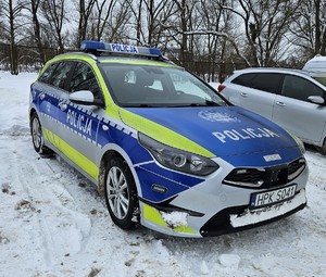 Oznakowany radiowóz stojący przodem. W tle ogrodzenie oraz  drzewa.