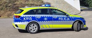 Na zdjęciu oznakowany radiowóz policyjny ustawiony bokiem. W tle widać betonowy most.