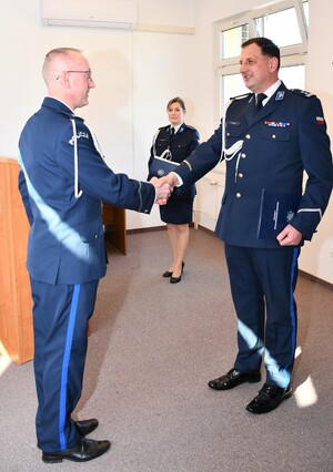 Na zdjęciu widać umundurowanych policjantów stojących naprzeciwko siebie, podających sobie prawe dłonie. Na drugim planie stoi umundurowana policjantka.