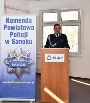 Na zdjęciu Komendant Powiatowy Policji w Sanoku mł. insp. Artur Kowalik stojący przy mównicy. Obok niego postawiony baner zawierający rysunek policyjnej odznaki oraz napis Komenda Powiatowa Policji w Sanoku.