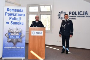 Na zdjęciu Zastępca komendanta Powiatowego Policji w Sanoku podinsp. Krzysztof Belczyk przemawiający przy mównicy. Obok niego stoi mło insp. Artur Kowalik, Komendant Powiatowy Policji w Sanoku. W tle okno.