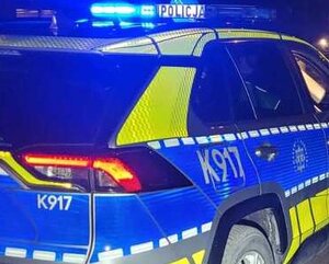 Na zdjęciu znajduje się oznakowany radiowóz policyjny z włączonymi światłami uprzywilejowania w kolorze niebieskim. Jest noc.