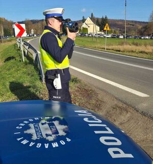 Na zdjęciu policjant z ruchu drogowego mierzący prędkość kierującym na drodze. W tle zaparkowane samochody oraz tereny zielone. Na pierwszym planie maska radiowozu policyjnego z napisem POLICJA oraz hasłem Pomagamy i chronimy.