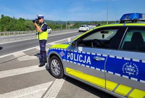 Umundurowany policjant używający ręcznego miernika prędkości. Obok oznakowany radiowóz policyjny. W tle droga oraz las.